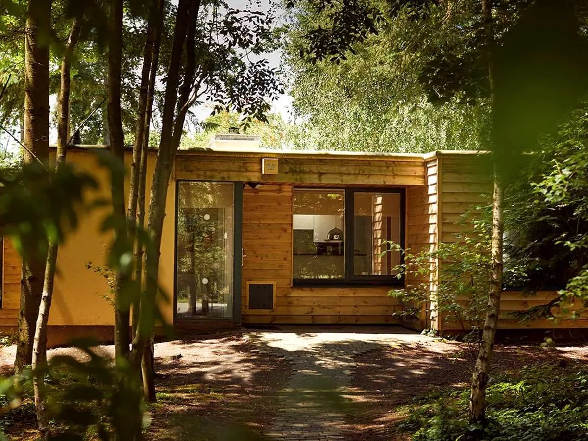 A single-storey timber lodge with full-height glass doors and a brick path to the entrance, surrounded by young trees.