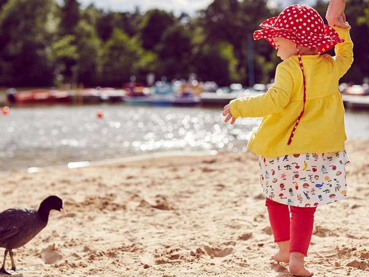 A toddler in a red sun hat and yellow cardigan walking on pale sand at the edge of the lake, with a moorhen nearby and boats moored beyond.