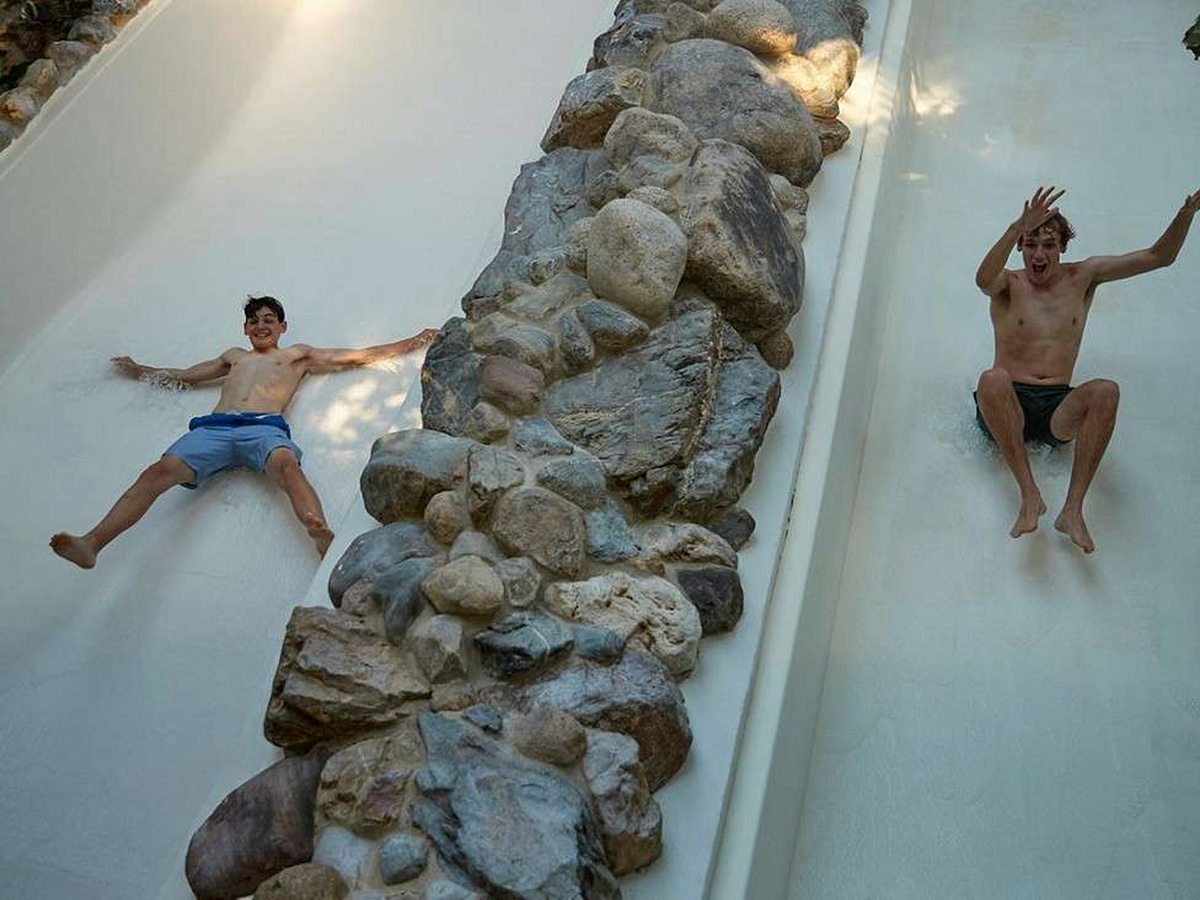 Two swimmers sliding down side-by-side white open flumes divided by a rock feature.