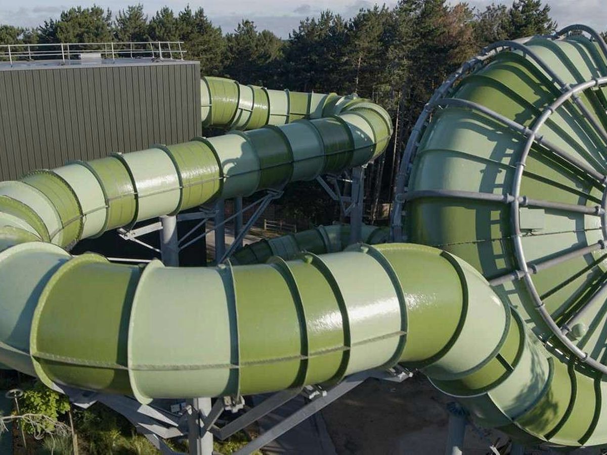 Pale green enclosed waterslide flumes coiling down the outside of the pool building against a background of pine trees.