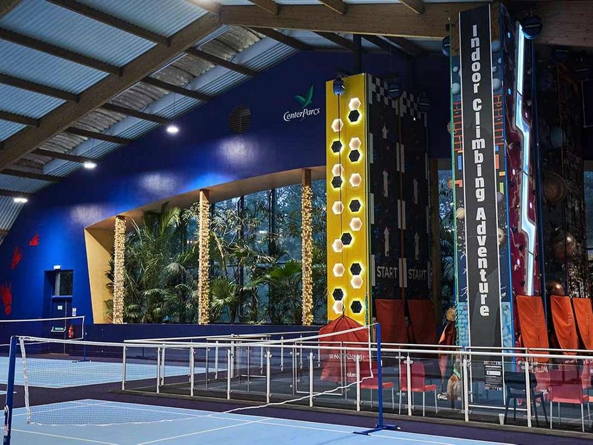 A view down the indoor sports hall, past a badminton net on a blue marked court, to the tall multi-lane Indoor Climbing Adventure wall at the far end.
