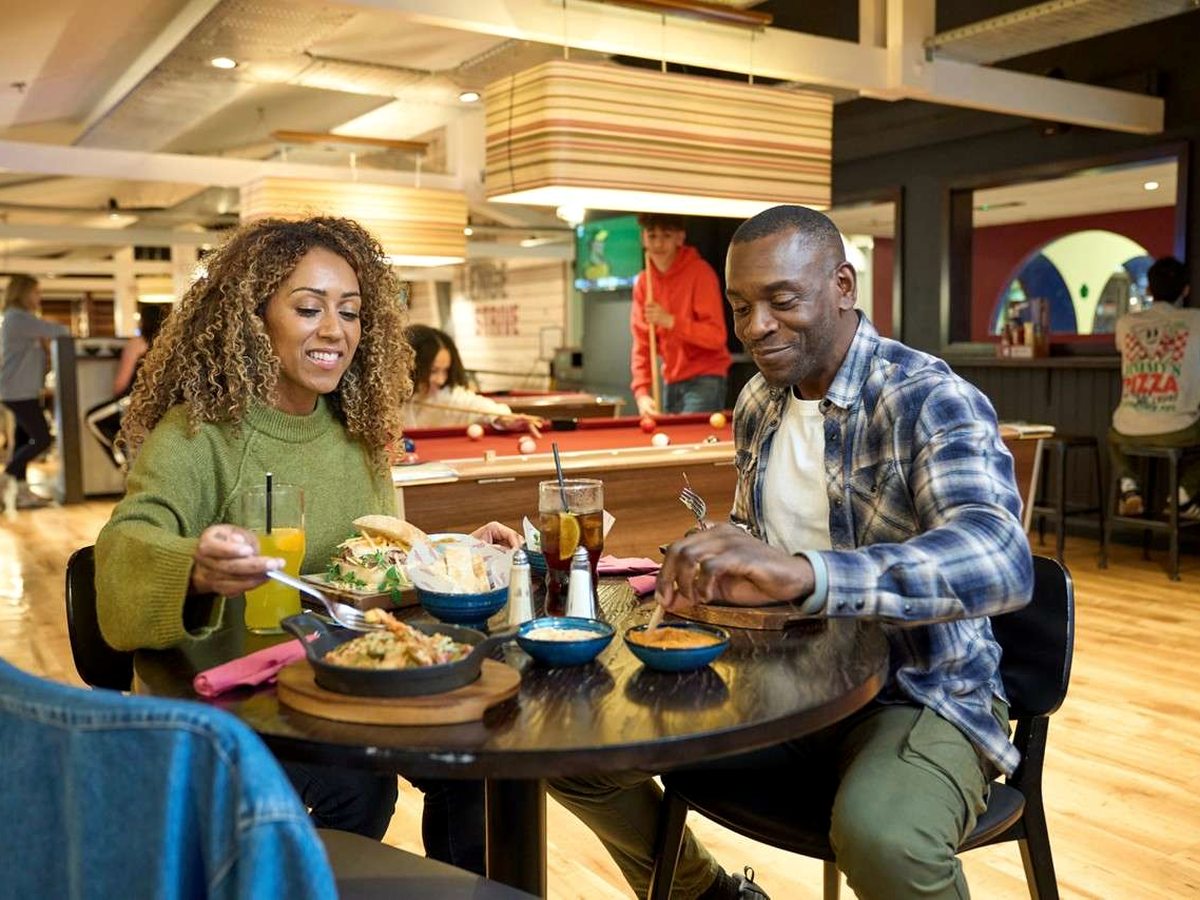 Two adults eating burgers and sharing plates at a round table in a wood-floored restaurant, with pool tables and a sports screen behind them.