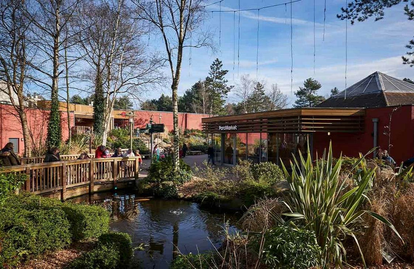 The village centre at Center Parcs Sherwood Forest, with the ParcMarket shop beside a planted pond and a timber boardwalk where families are sitting out in the sun.