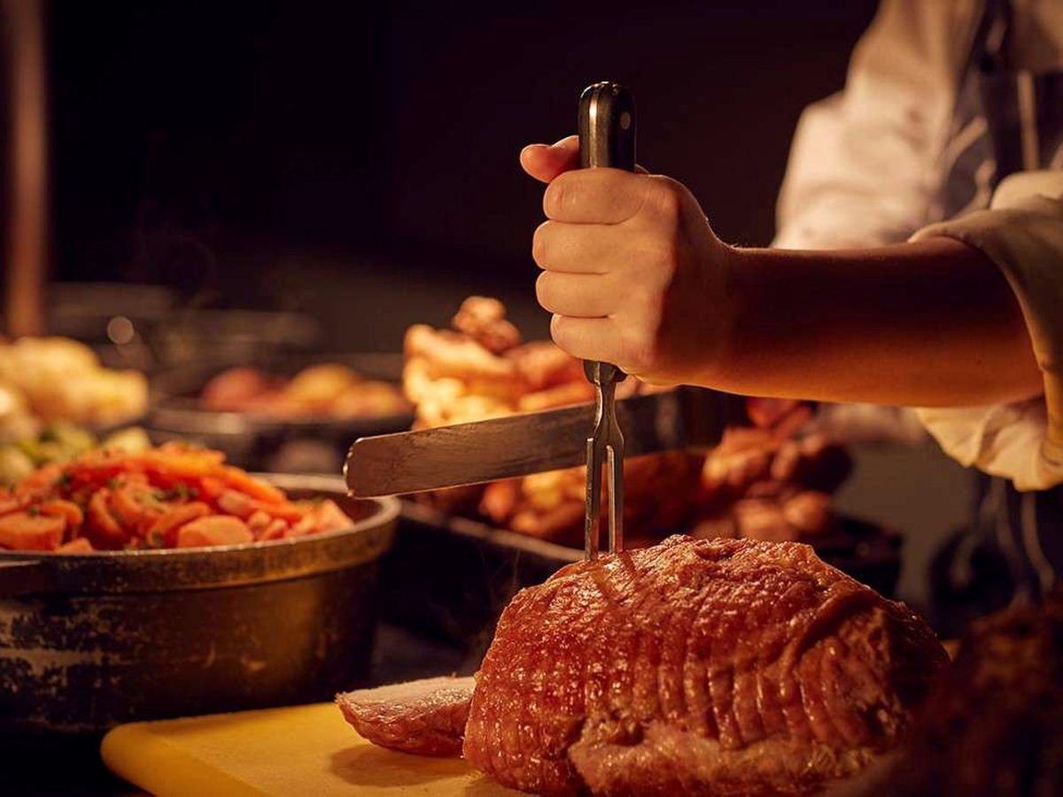 A chef in whites carving a joint of roast meat with a knife and fork at a hot counter, with trays of roast vegetables and Yorkshire puddings behind.