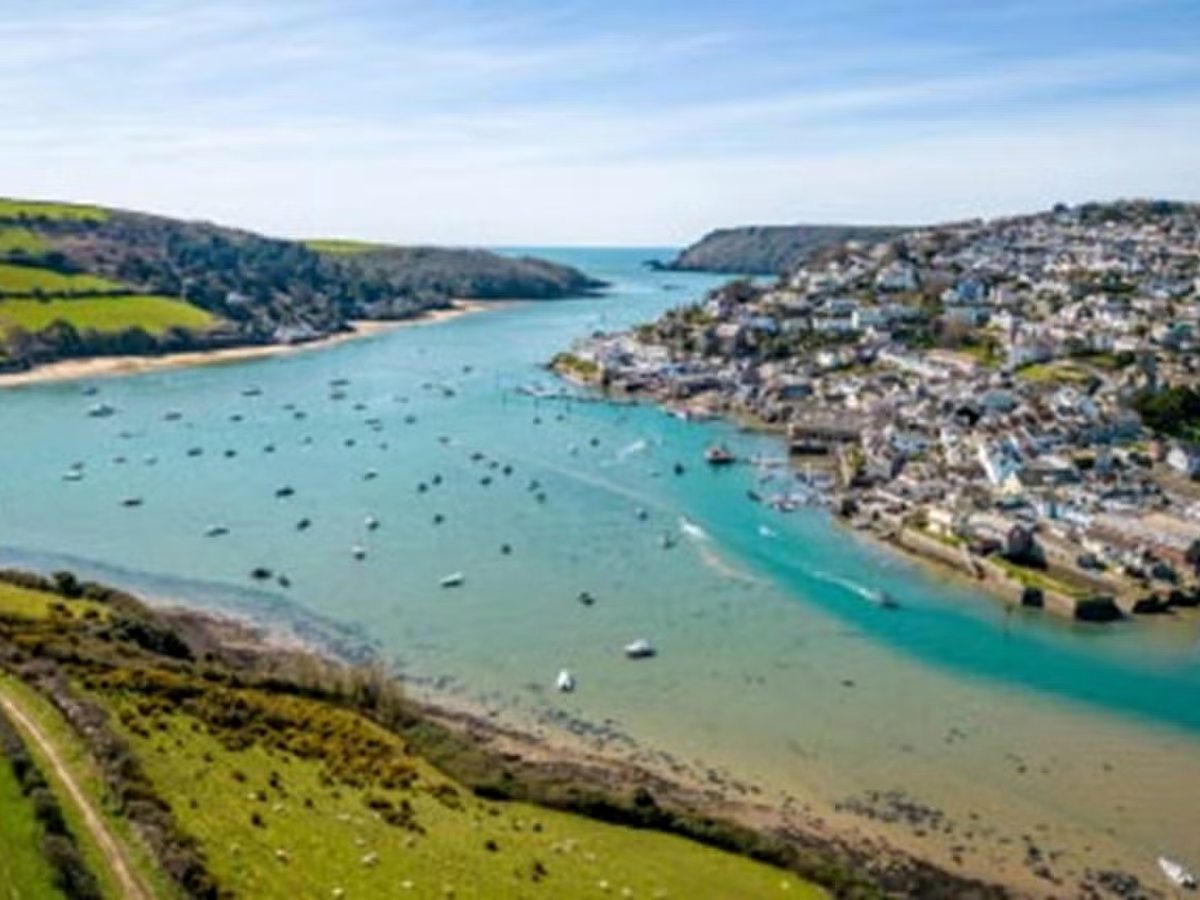 Aerial view of a turquoise tidal estuary dotted with moored boats, with a waterside town spread along the far shore and green hills beyond.