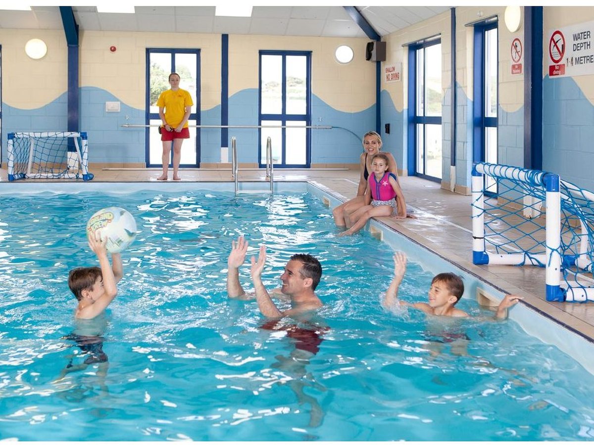 Children playing with a ball in a shallow indoor swimming pool with pale blue wave-painted walls, tall windows and a lifeguard at the poolside.
