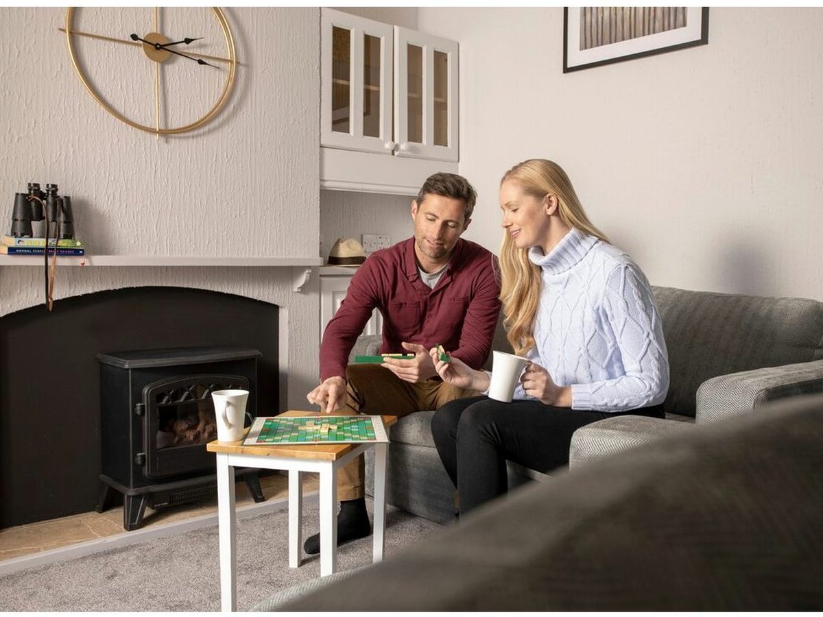 A carpeted open-plan living area with a grey corner sofa, an electric stove set in a white surround, overhead cabinets and a large wall clock.