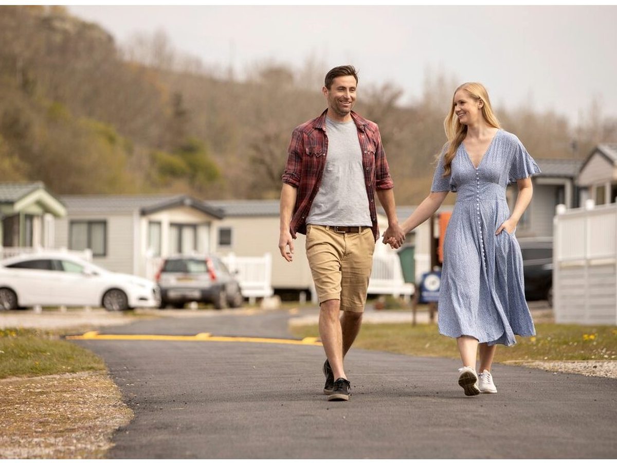 A paved internal road running between rows of white static caravans, with parked cars, a couple walking and a wooded hillside behind.