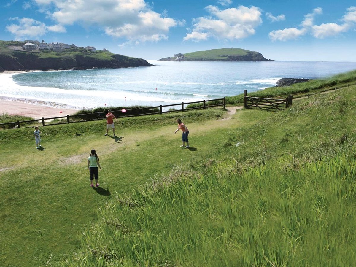 Children playing on a sloping mown lawn above the beach, with a surf-lined bay, a green headland and an offshore island in the distance.