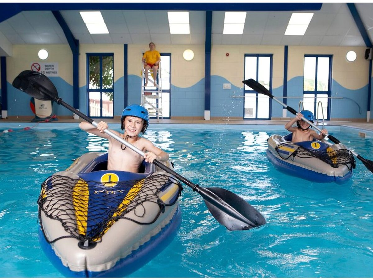 Children in buoyancy vests and helmets paddling inflatable kayaks across the indoor pool, watched by an instructor at the poolside.
