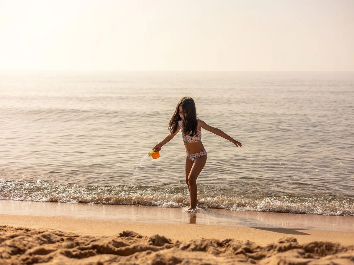 A young child in a swimsuit pouring water from a bucket while standing ankle-deep in the calm shallows at the sandy shoreline in soft light.