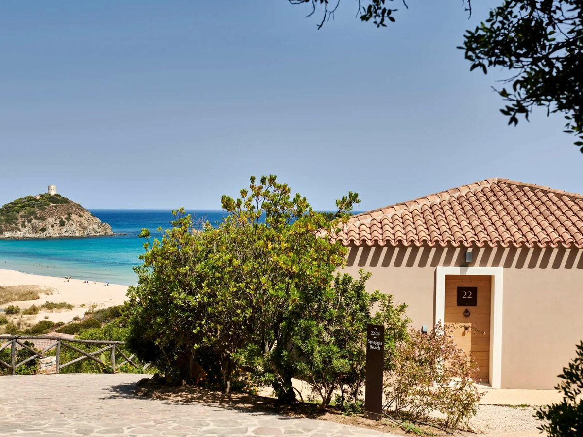 A single-storey guest cottage with a terracotta-tiled roof set among green Mediterranean shrubs, with a sandy beach and the Chia Tower headland behind under a clear blue sky.