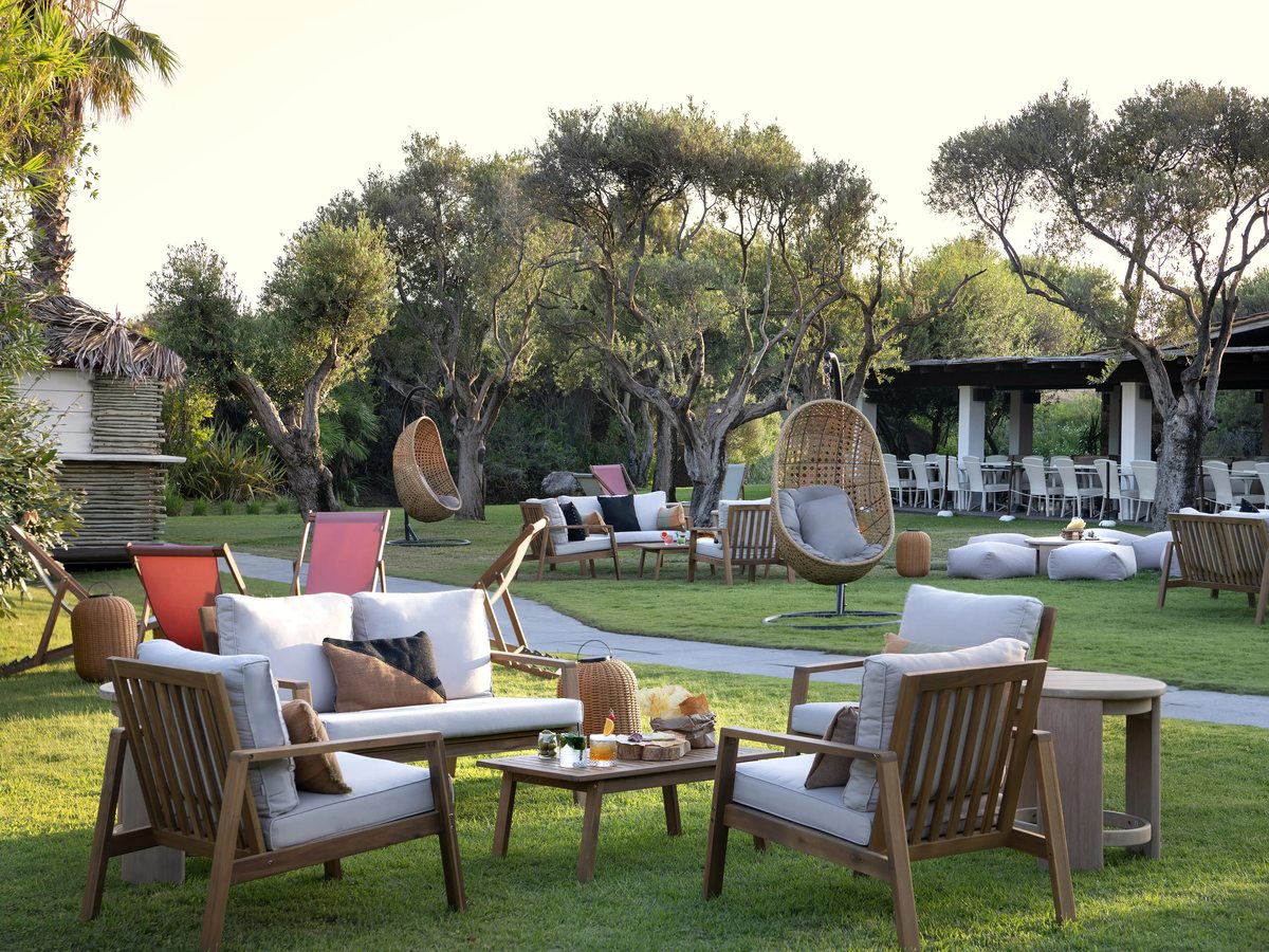 An outdoor garden seating area with wooden-framed sofas, cushioned hanging chairs and low tables set on a lawn among olive trees, with a covered dining pavilion beyond.