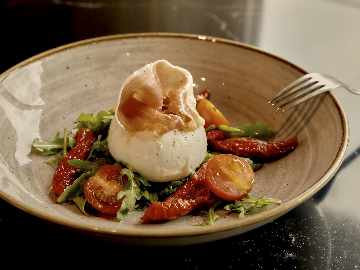 A ball of burrata on a plate with cured ham, cherry tomatoes, sun-dried tomatoes and salad leaves.