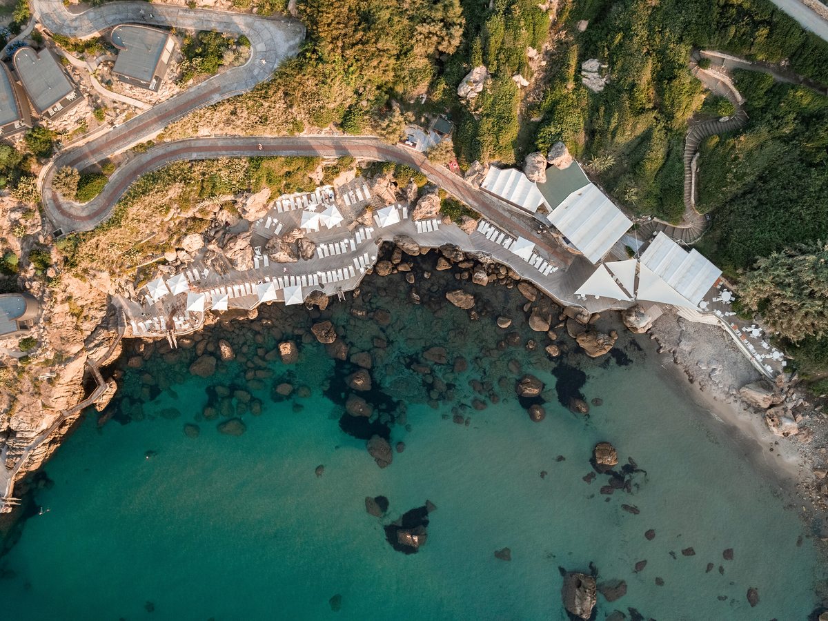 Aerial view of a rocky cove with clear turquoise water, wooden sun decks and rows of white parasols along the shore.