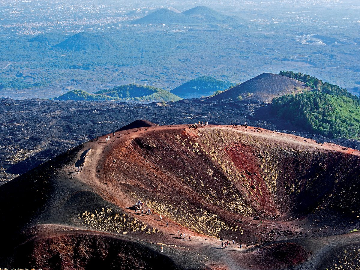 The red-and-black volcanic crater and lava fields of Mount Etna with tiny hikers on a winding path and forested cones beyond.