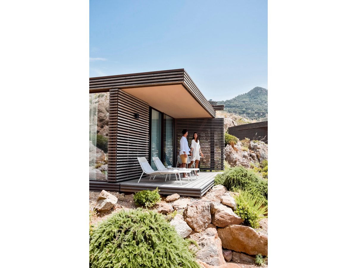 A timber-slatted Villetta bungalow with a couple standing on its private sun-deck terrace on a rocky, planted slope.