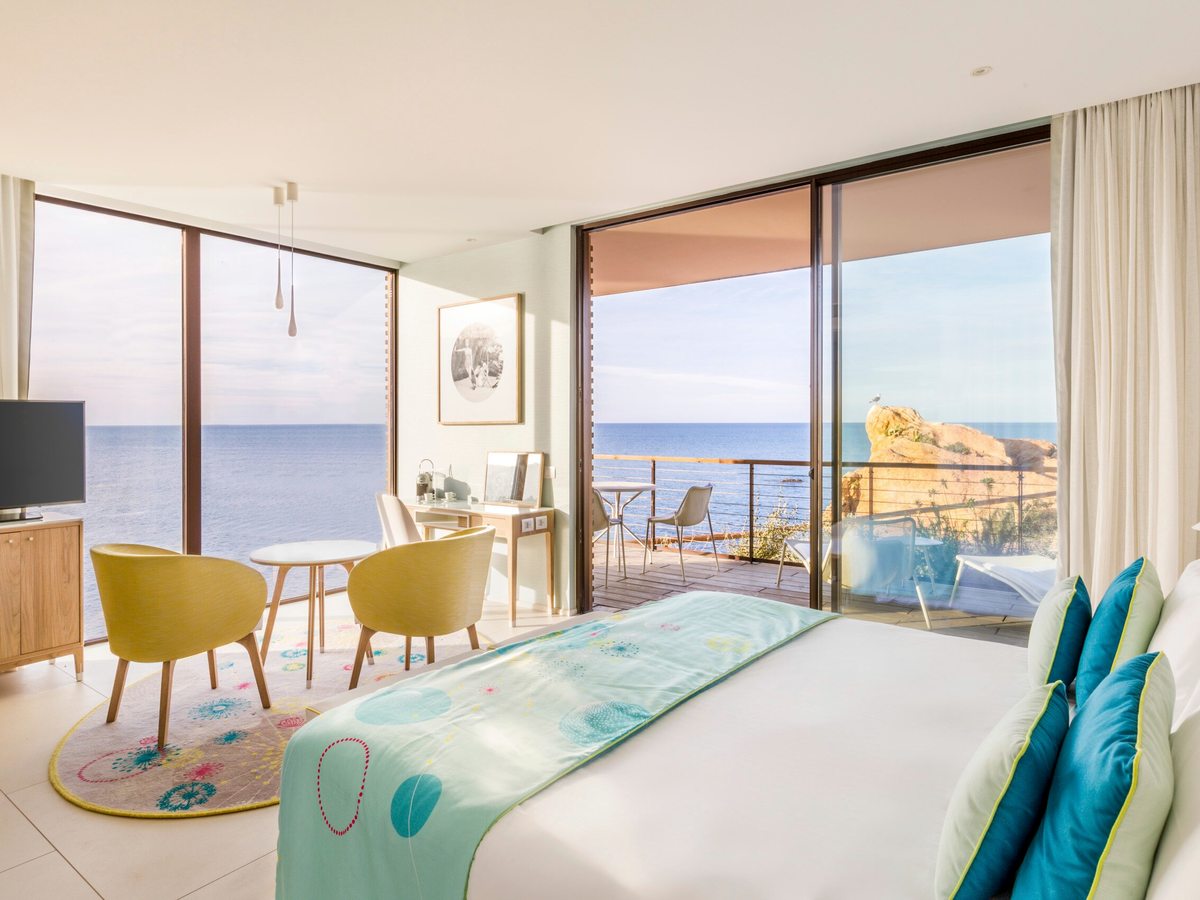 A bright bedroom with a double bed, yellow tub chairs and a writing desk, with floor-to-ceiling windows opening onto a terrace above the sea.
