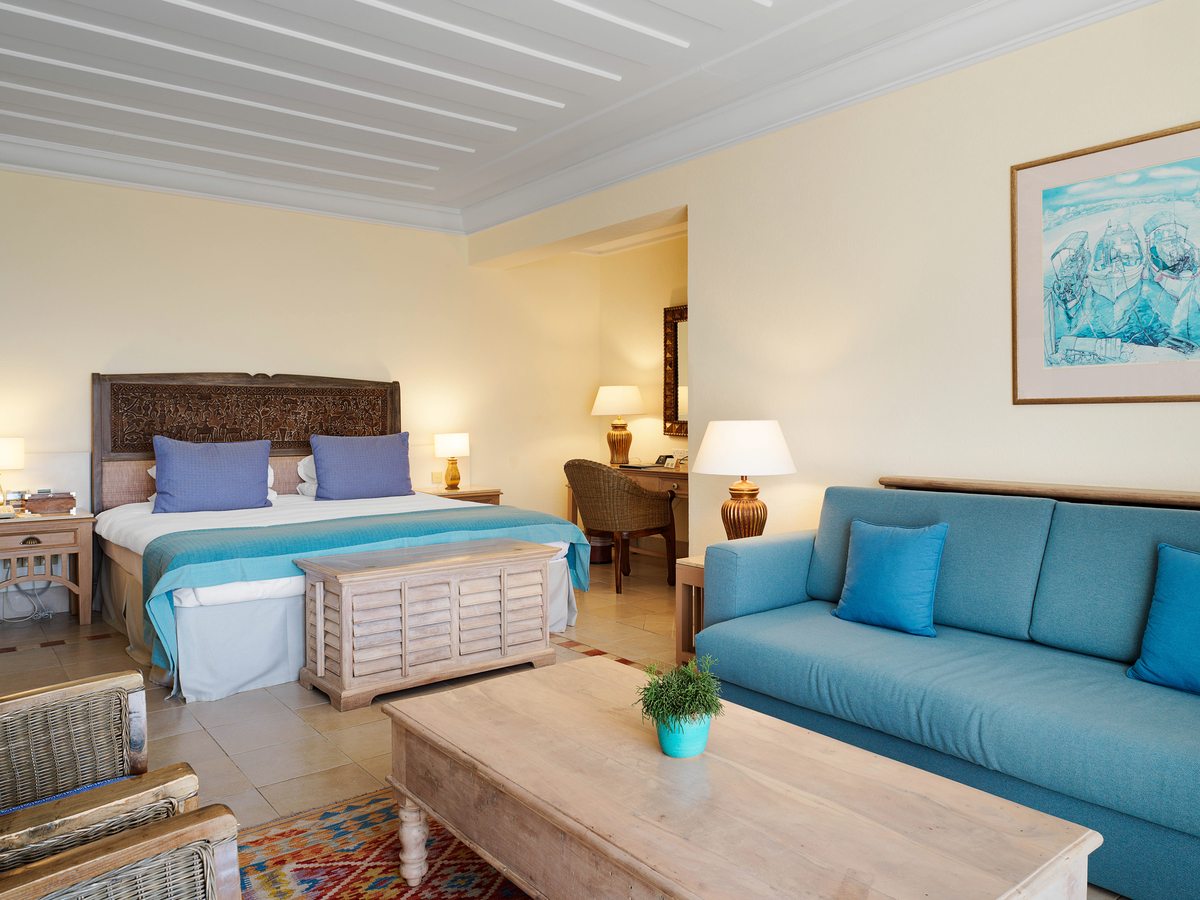 An open-plan suite with a double bed, a blue sofa, a wooden coffee table and coastal artwork on tiled floors.