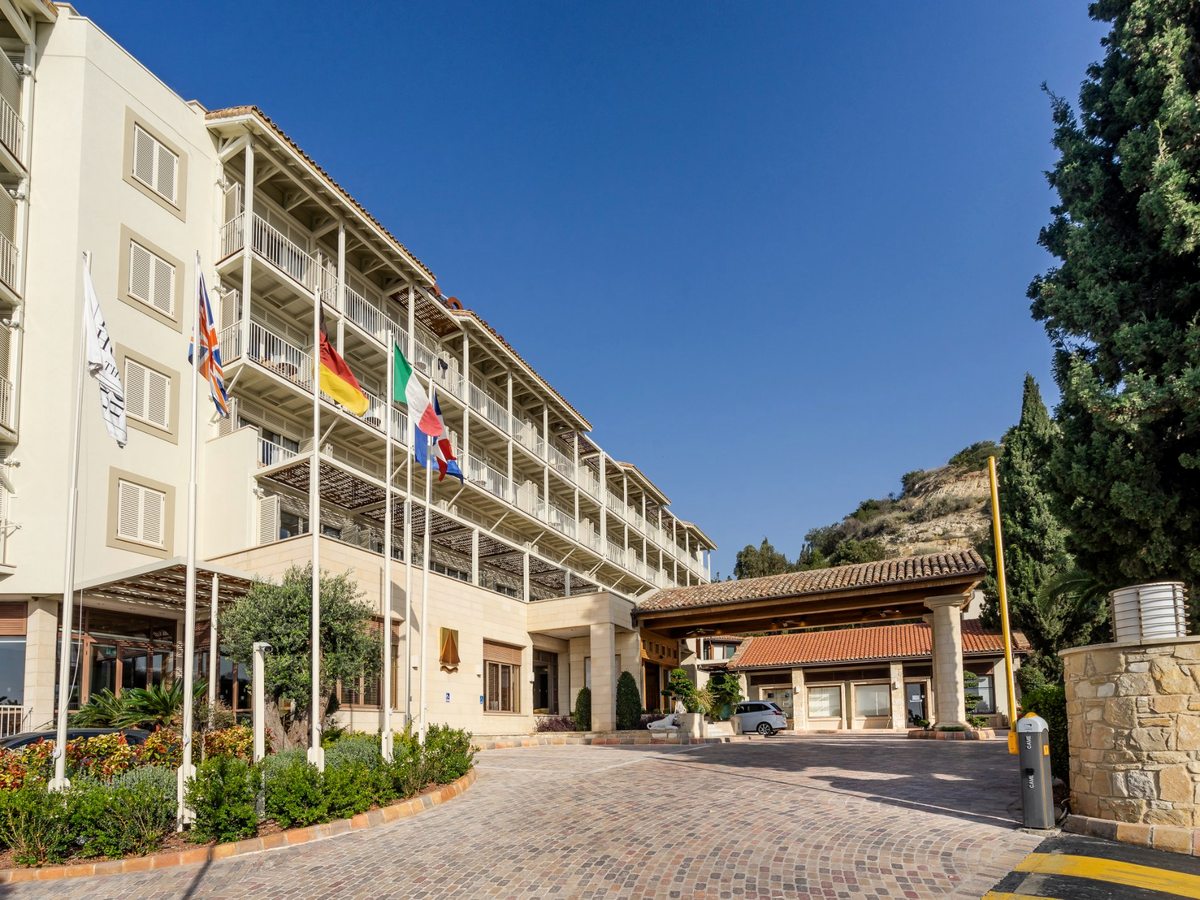 The main hotel building with a covered stone entrance porch, a paved forecourt and a row of national flags.