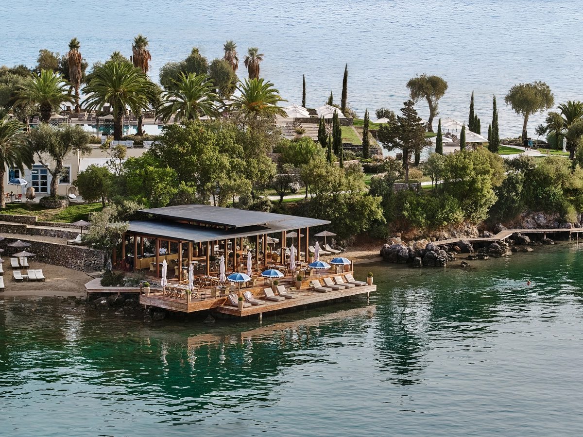Open-sided waterfront restaurant on a wooden deck built out over a calm green cove, with dining tables, sun loungers and blue parasols, backed by palms and cypress trees.