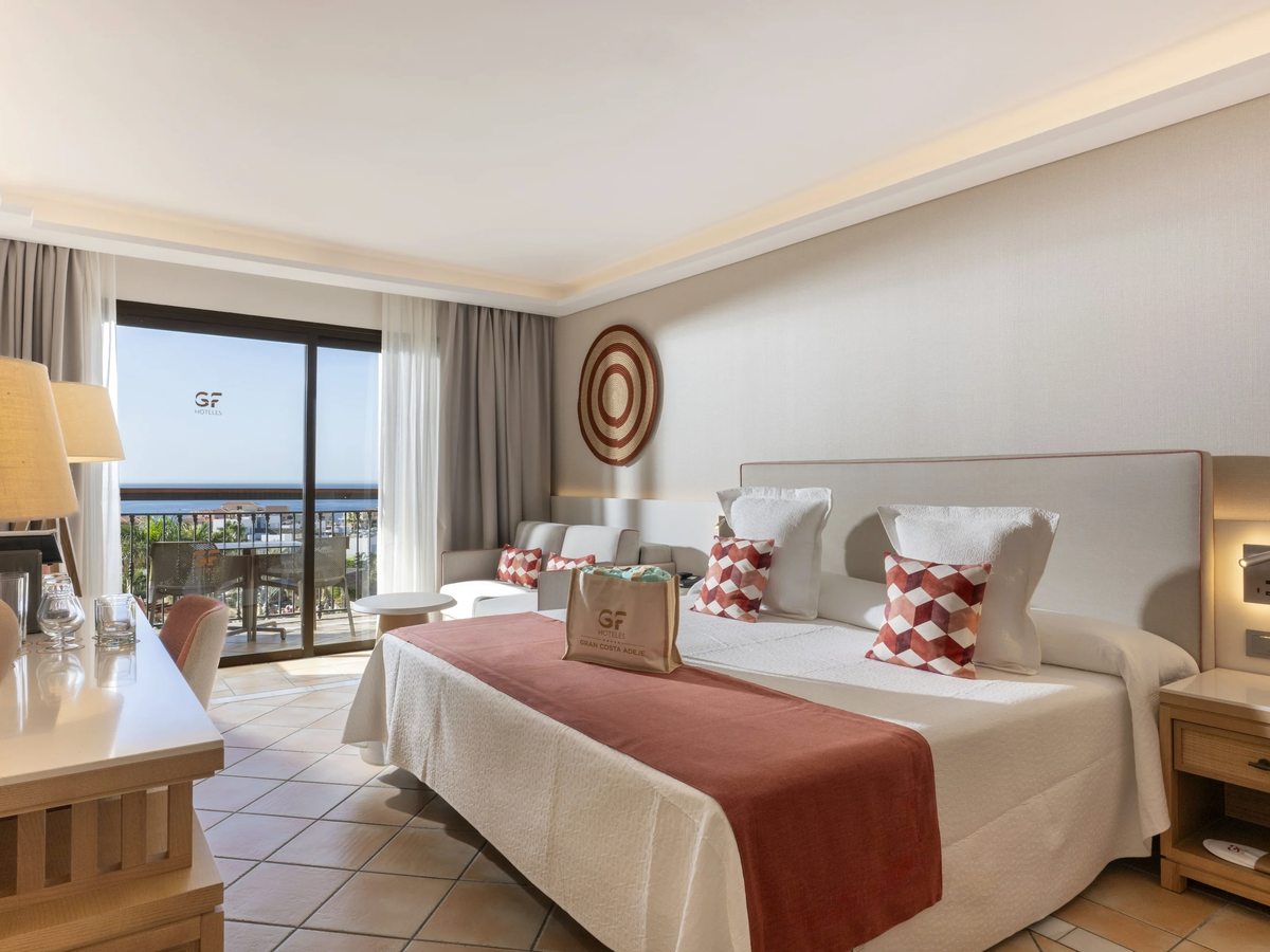 A double room with a large bed, a rust-red bed runner, a writing desk and sliding doors onto a sea-view balcony.