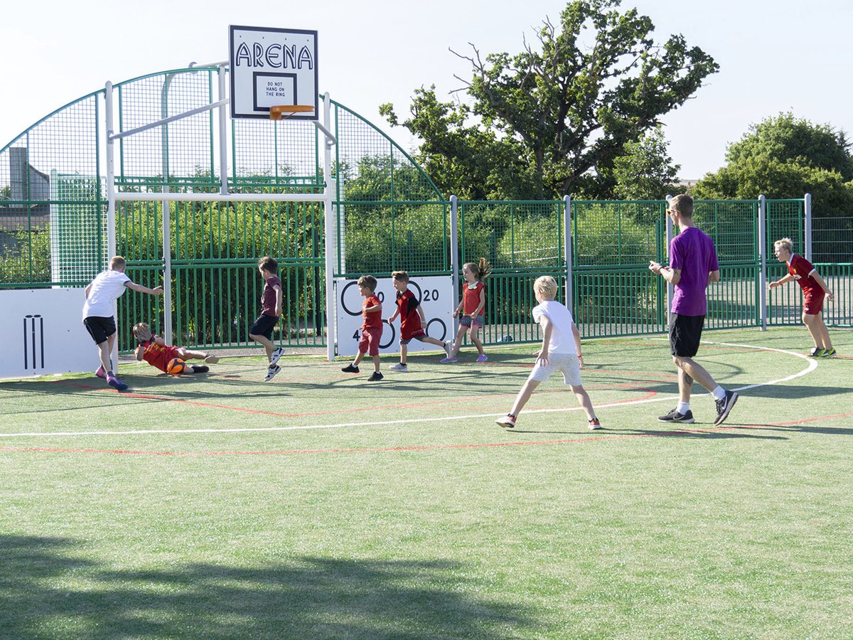 Children playing football on a green all-weather sports court with an Arena basketball hoop, trees behind