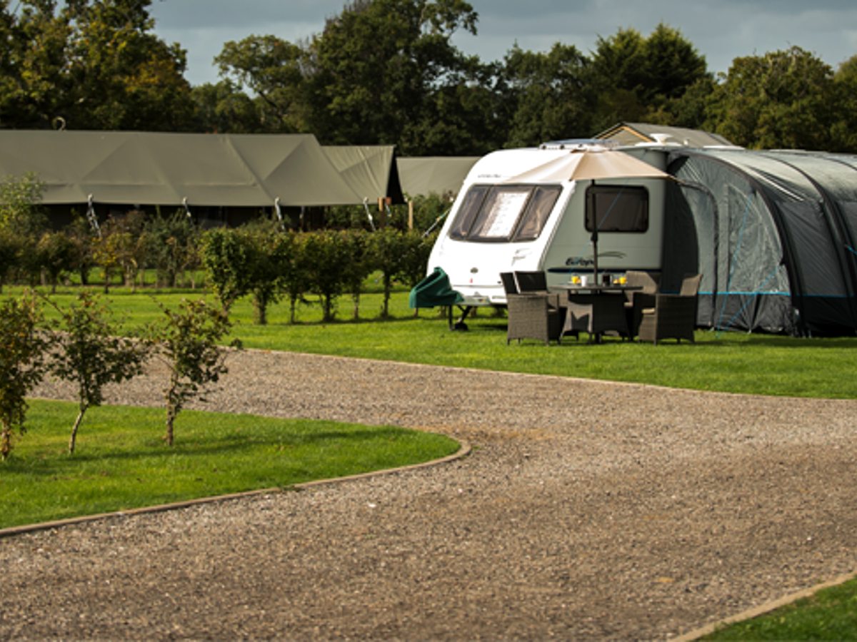 A touring caravan with an awning on a grass pitch, with safari tents along the treeline behind