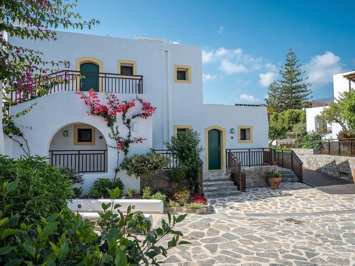 Whitewashed two-storey resort buildings with green shutters and bright pink bougainvillea above a stone-paved path