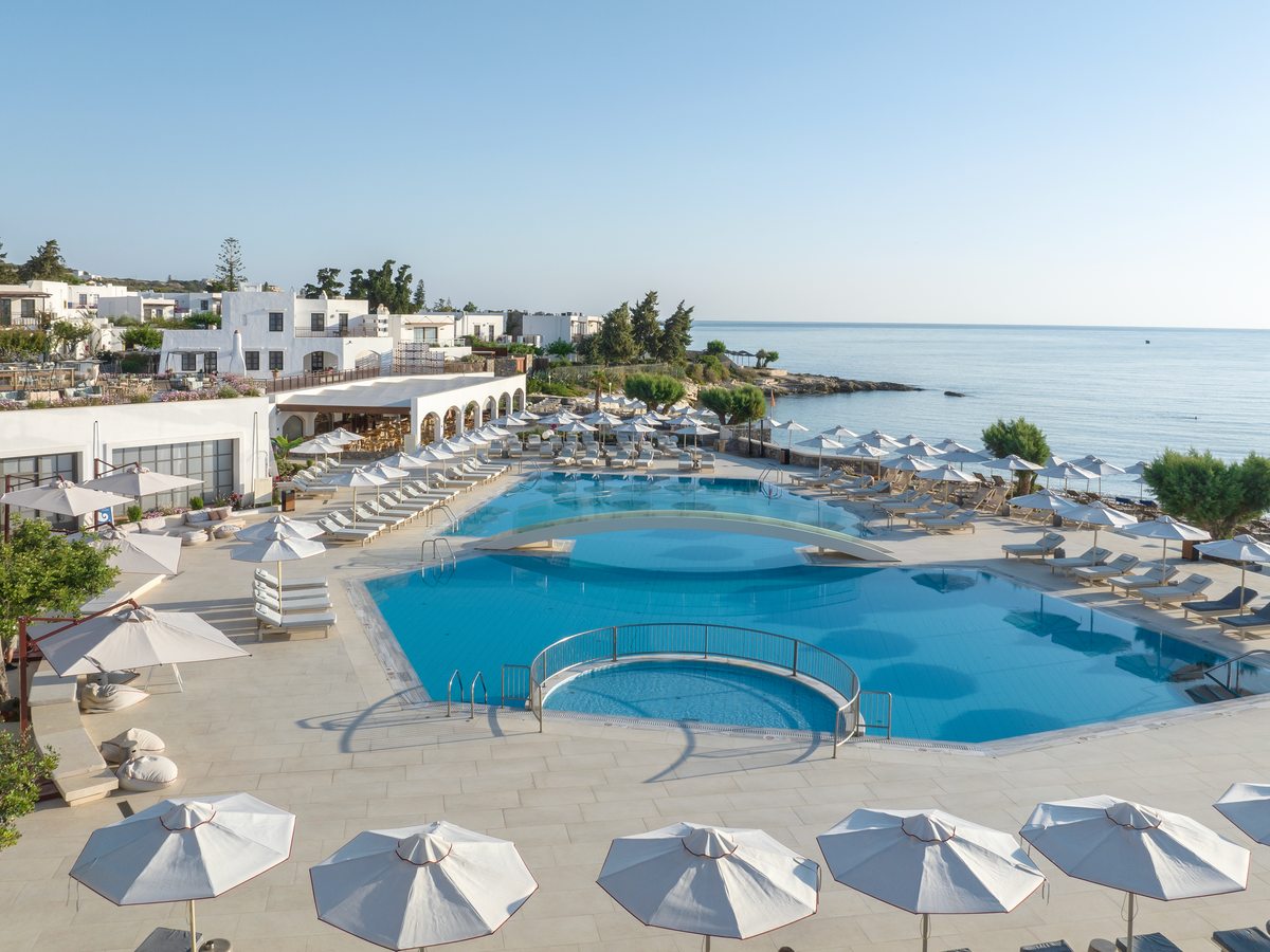 Two large freshwater pools ringed by white umbrellas and loungers, with the whitewashed resort and the sea behind