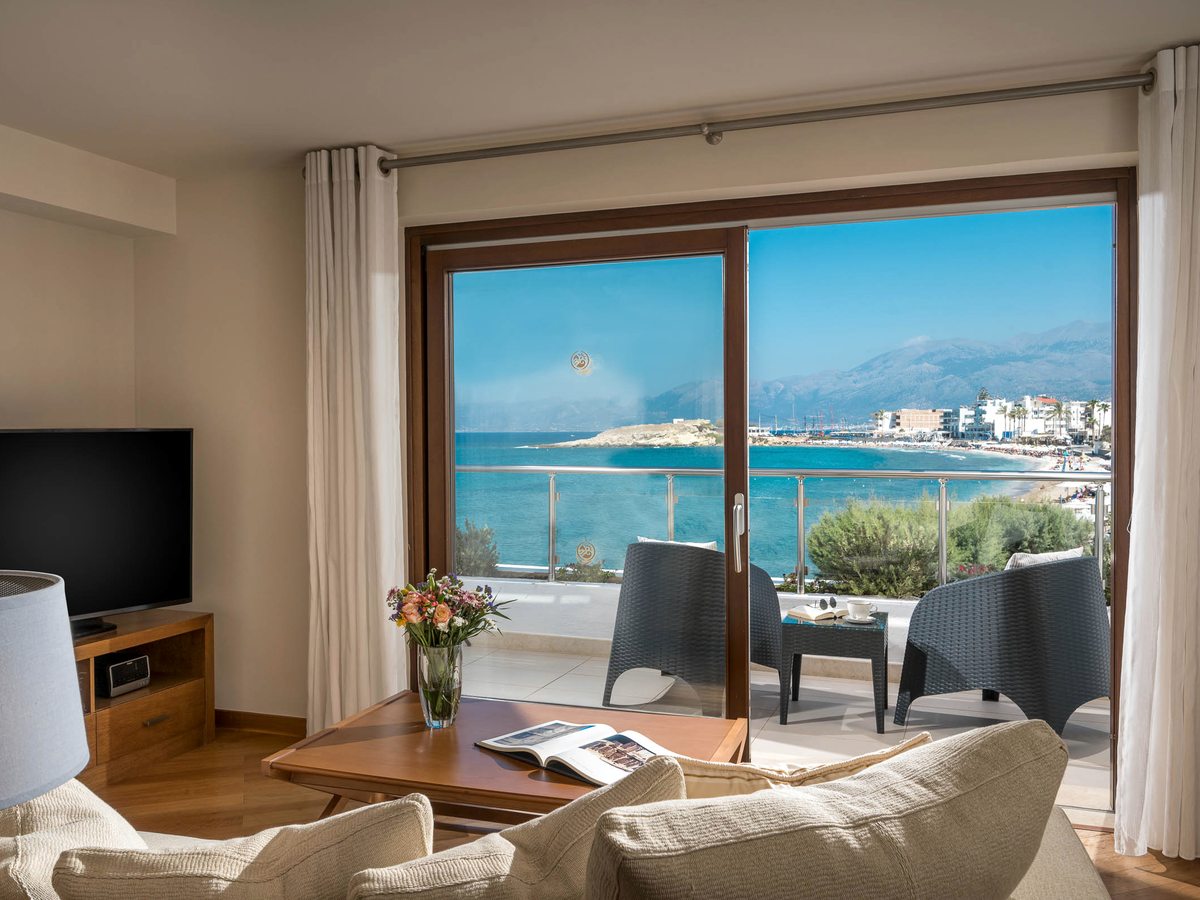 A suite sitting room with cream sofas and armchairs opening to a balcony that looks over the bay towards Hersonissos