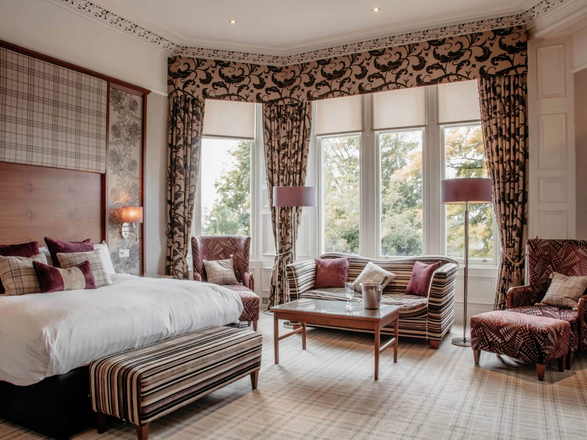 A hotel double bedroom with a large bed, tartan cushions, two armchairs and a coffee table by bay windows with patterned curtains.