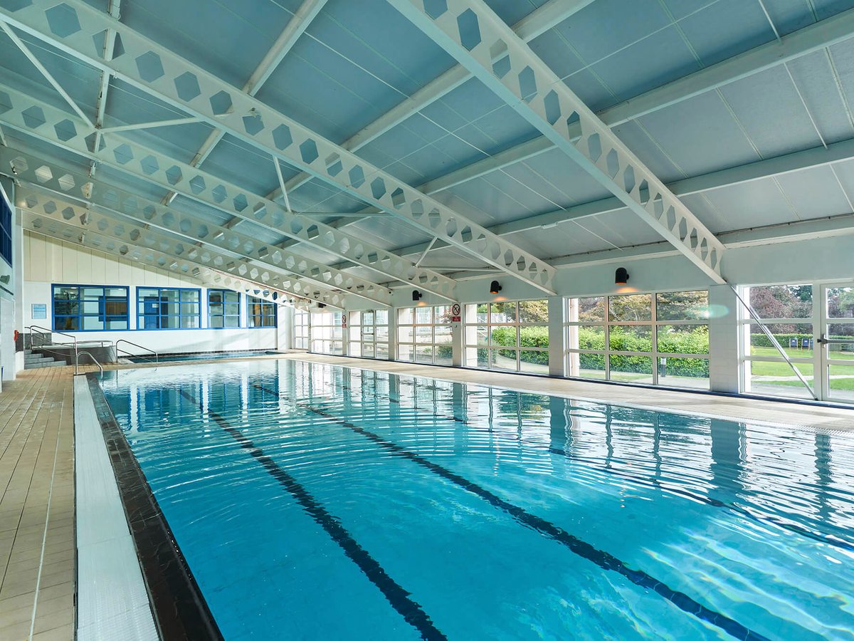 An indoor swimming pool with marked lanes under a white pitched roof, with large windows onto greenery along one side.