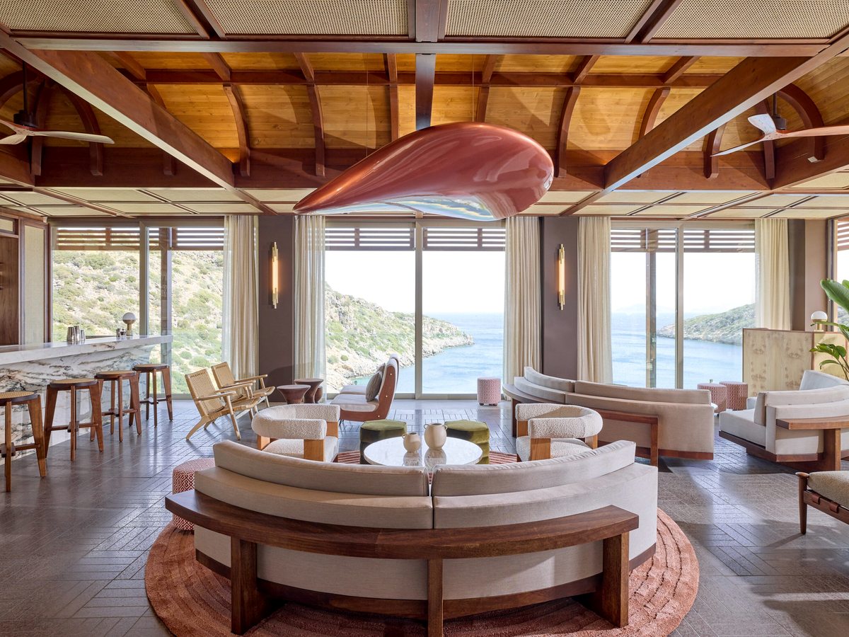 A bar lounge with floor-to-ceiling windows over the sea, a curved banquette and low seating beneath a sculptural ceiling.