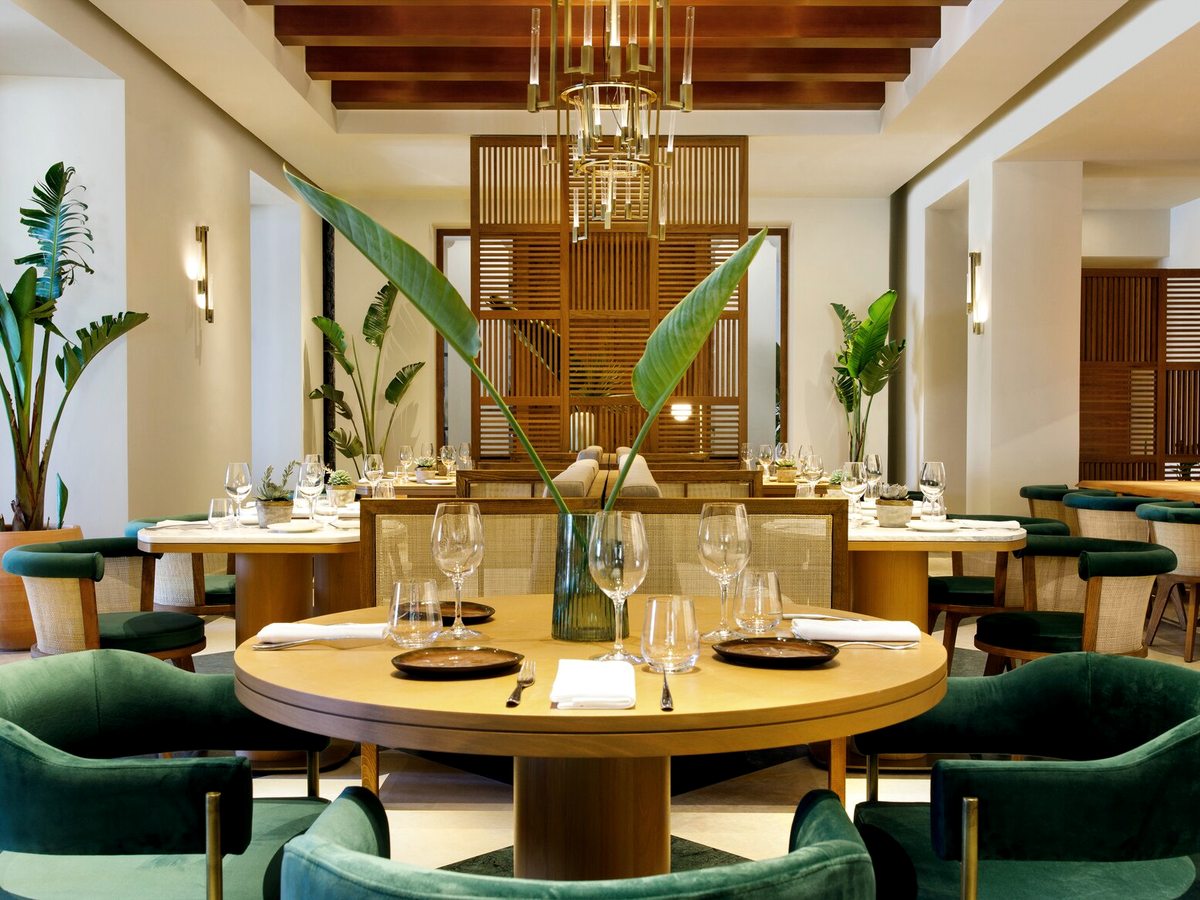 A restaurant interior with round tables, green velvet armchairs, tall tropical plants and a timber-beamed ceiling.