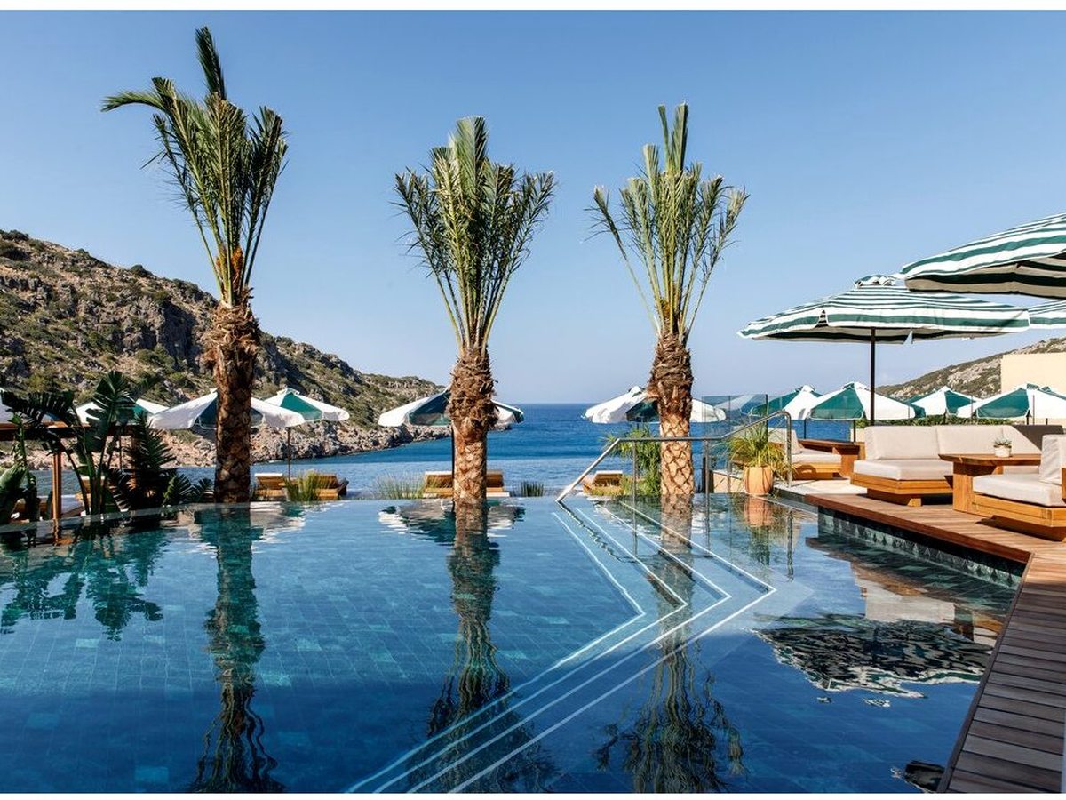 A still pool lined with tall palms, cushioned daybeds and green-striped parasols on a wooden deck above the sea.