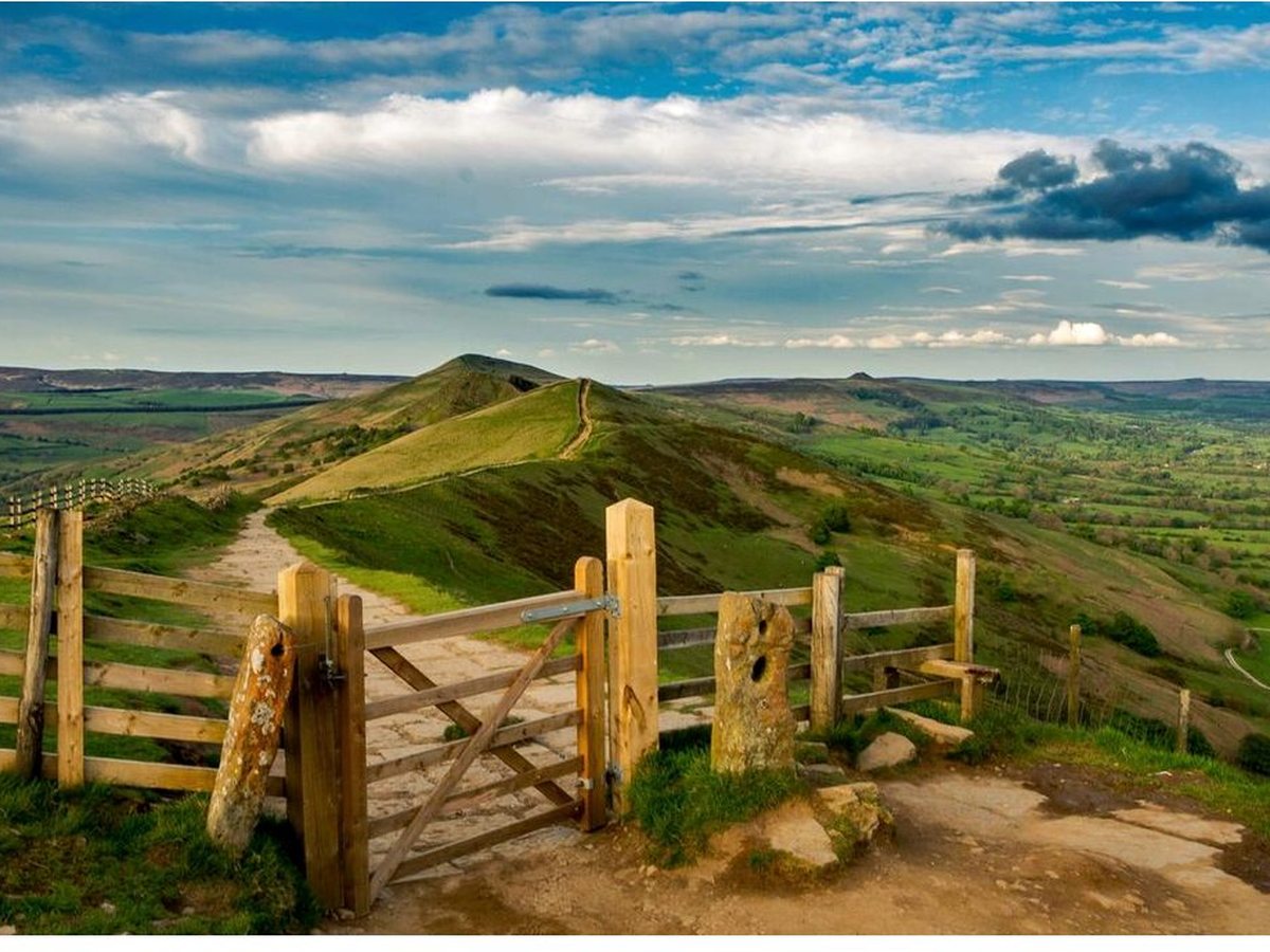 A grassy hilltop viewpoint in the Peak District with a wooden gate, a fenced footpath and rolling fields beyond.