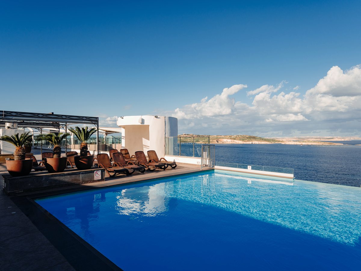 A rooftop infinity pool with wooden sun loungers and potted palms, looking out over the sea to the coastline beyond under a bright blue sky.