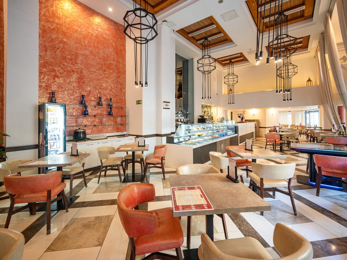 A café interior with a glass cake-and-pastry display counter, a service counter, tub chairs at café tables and geometric pendant lights under a high ceiling.