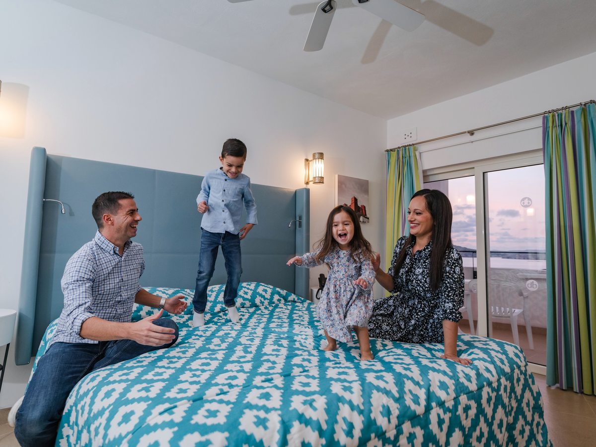 A bright room with a double bed dressed in turquoise patterned covers, two children standing on the bed with their parents beside a balcony door.