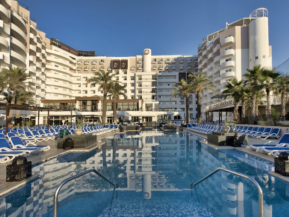 A large rectangular pool ringed with blue and white sun loungers and palms, the curved white multi-storey hotel with its rooftop db sign rising behind.