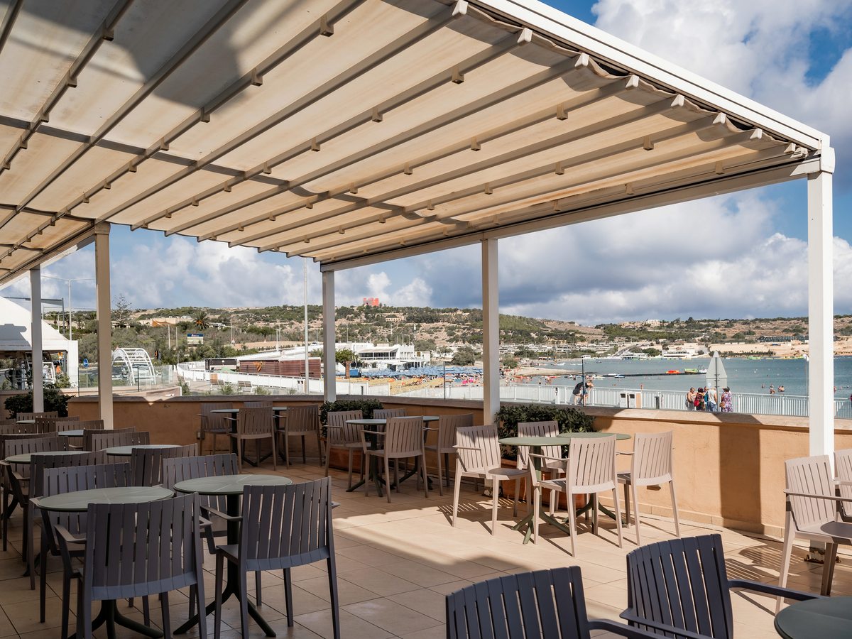A covered terrace of tables and chairs under a retractable canopy, looking out over Mellieħa Bay, the sandy beach and moored boats.