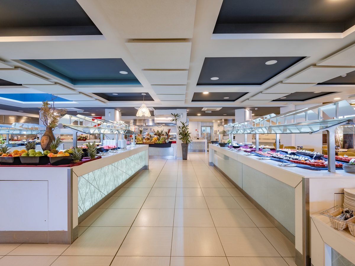 A large self-service buffet restaurant with island serving counters under glass guards, stacks of plates and a wide tiled aisle between dining tables.