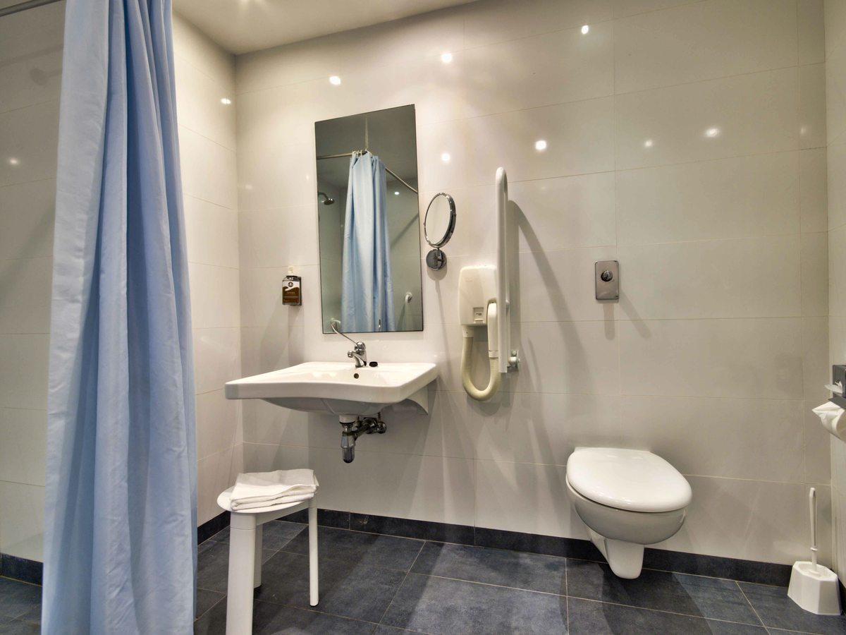 An accessible bathroom with a step-free roll-in shower, a fold-down shower seat and handheld shower, grab rails, a wall-mounted basin and a wall-hung toilet.