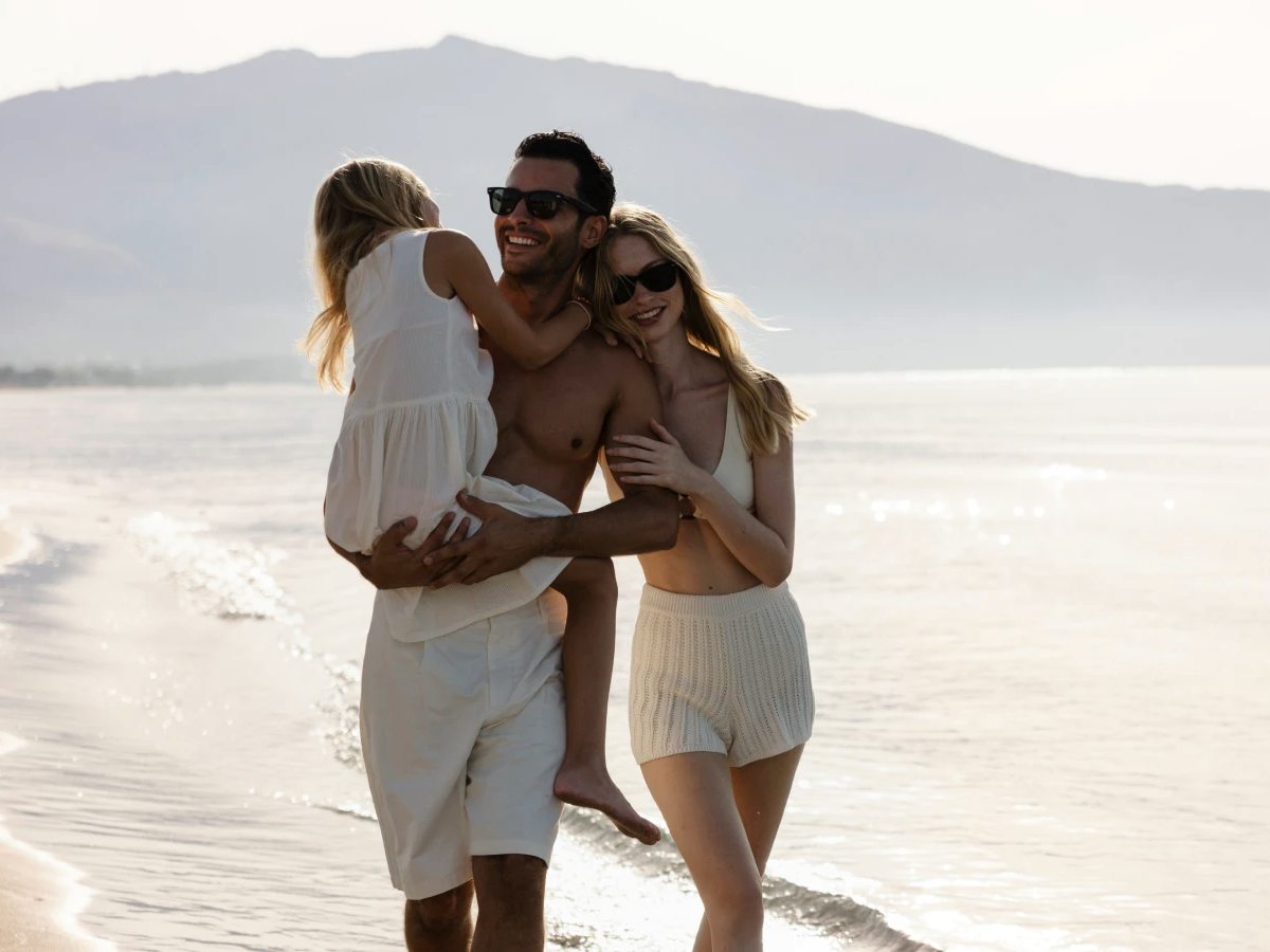 Family walking along the sandy shoreline at the water's edge, with calm sea and hazy mountains behind.