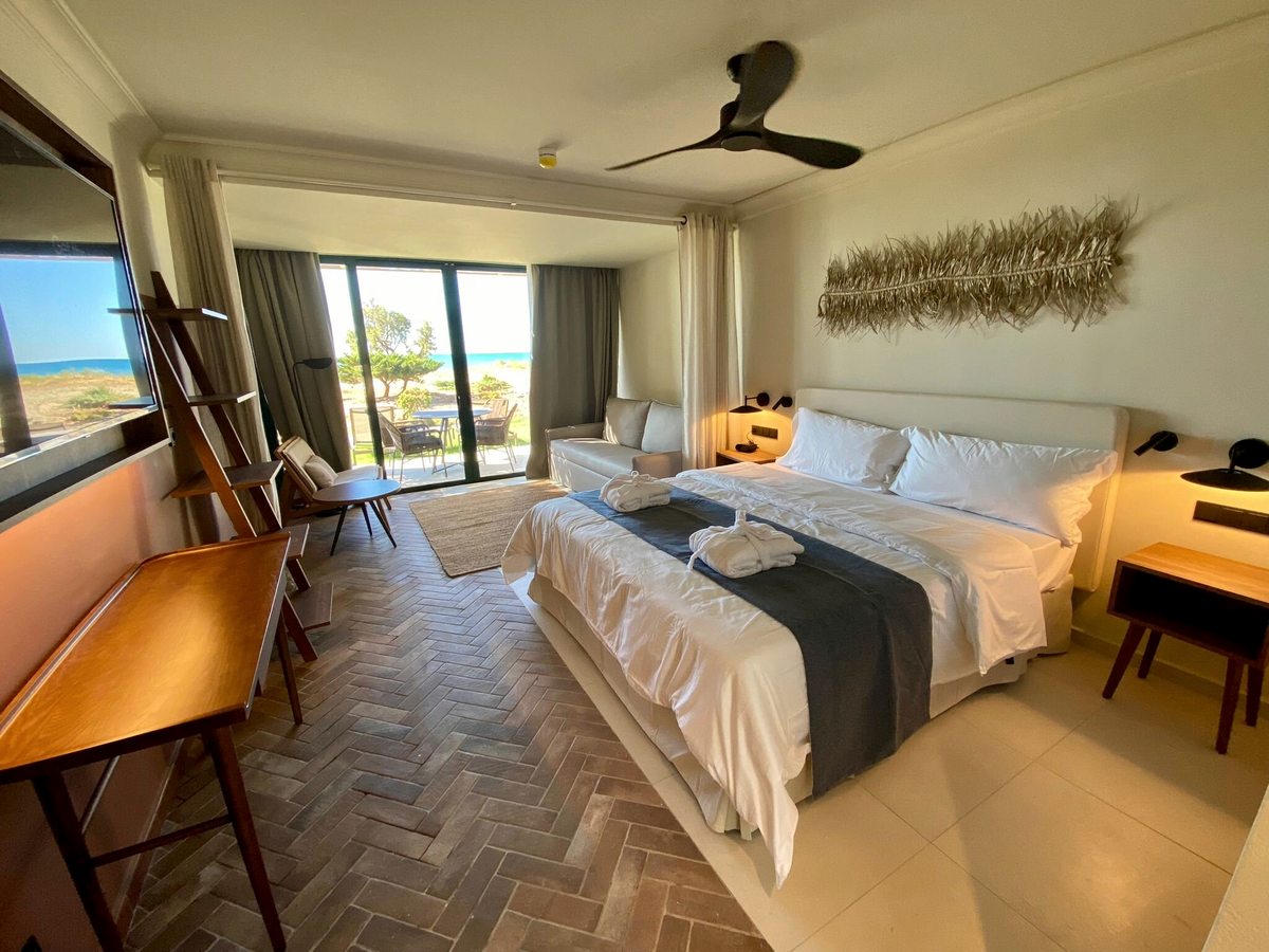 Double room with a large bed facing sliding glass doors to a furnished terrace and a coastal view, with a wall-mounted TV and a wooden desk.