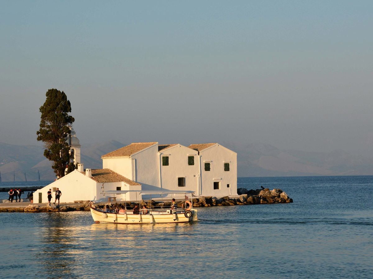 The small whitewashed Vlacherna Monastery on an islet linked by a causeway, with a cypress tree and a wooden excursion boat on calm water.