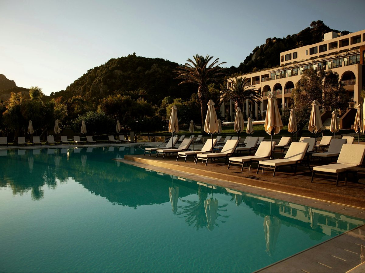 Large turquoise pool edged with cushioned sun loungers and closed parasols, the tiered arched resort building and wooded hillside behind.