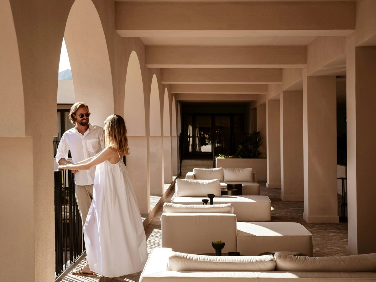 Covered terrace lined with repeating arches and columns, cream sofas and low tables, with a couple seated at the far end.