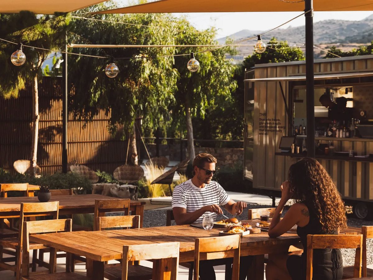 Wooden picnic tables under string lights beside a container-kitchen bar, with a couple eating and a hillside behind.
